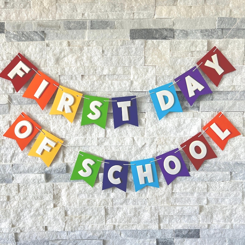 Puede incluir: Una colorida pancarta con las palabras "FIRST DAY OF SCHOOL" en letras blancas. Cada palabra est&aacute; en una bandera de bander&iacute;n de colores del arco&iacute;ris, unidas por una cuerda. El fondo es una pared de piedra gris claro.