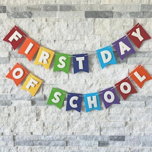 Puede incluir: Una colorida pancarta con las palabras "FIRST DAY OF SCHOOL" colgada en una pared de ladrillos blancos. La pancarta presenta banderas individuales en colores del arco&iacute;ris, con letras blancas. Una decoraci&oacute;n festiva.