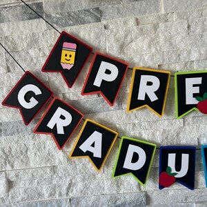 K&ouml;nnte beinhalten: Ein farbenfrohes Filzbanner mit den Worten "Pre-Grad" in wei&szlig;en Buchstaben auf schwarzen Filzdreiecken. Jedes Dreieck hat einen andersfarbigen Rand, darunter Rot, Orange, Gelb, Gr&uuml;n, Blau und Lila. Das Banner ist mit einem Bleistift und einem Apfel verziert.