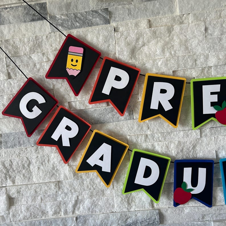 K&ouml;nnte beinhalten: Ein farbenfrohes Filzbanner mit den Worten "Pre-Grad" in wei&szlig;en Buchstaben auf schwarzen Filzdreiecken. Jedes Dreieck hat einen andersfarbigen Rand, darunter Rot, Orange, Gelb, Gr&uuml;n, Blau und Lila. Das Banner ist mit einem Bleistift und einem Apfel verziert.