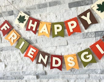 May include: A festive banner with the words "HAPPY FRIENDS GIVING" in white capital letters. The pennants are in fall colors: orange, red, brown, and green. Each pennant is shaped like a flag and strung together with a thin string. Some pennants have a green leaf.
