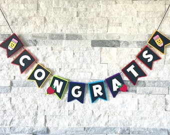 May include: A colourful banner spelling "CONGRATS" with black pennants, white letters, and pencil and apple accents. The banner is strung on a black string and is displayed against a textured white brick wall.