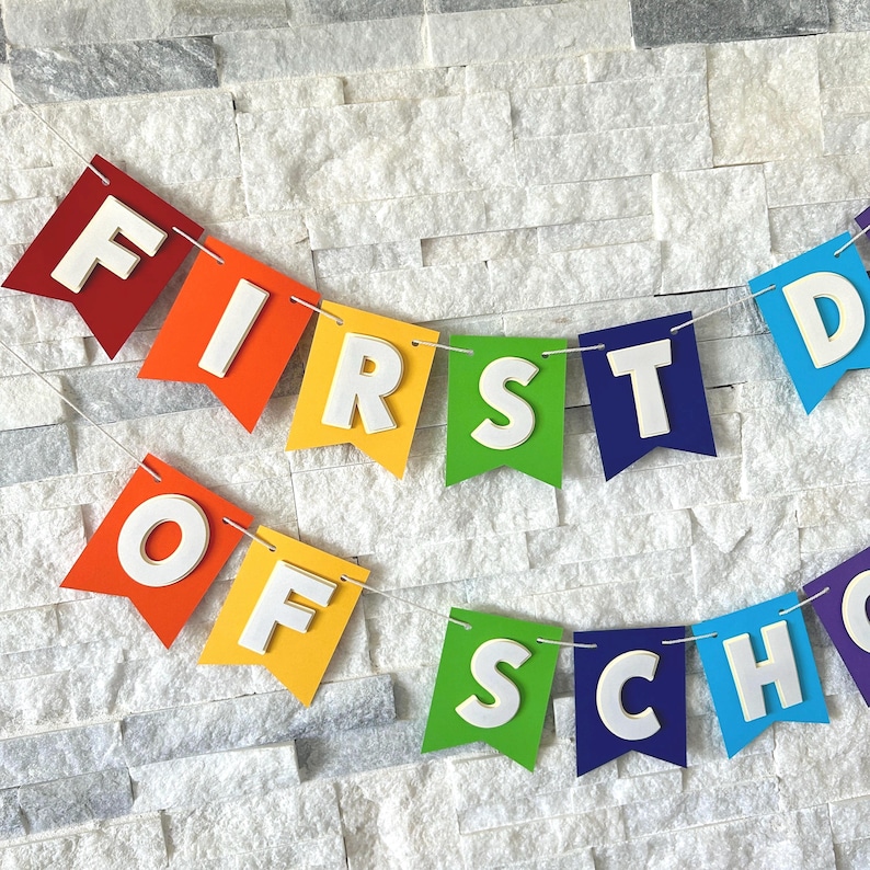 Puede incluir: Una colorida guirnalda con las palabras "FIRST DAY OF SCHOOL" ensartadas en una cuerda blanca. Cada bandera es de un color diferente, con letras blancas en 3D. El fondo es una pared de ladrillos blancos.