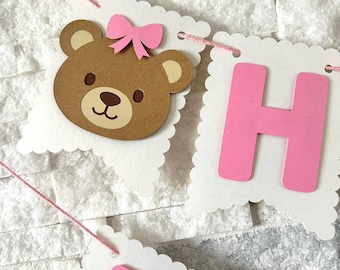 May include: A white banner with pink and brown cutouts. The banner says "Happy" with a brown teddy bear with a pink bow on its head.