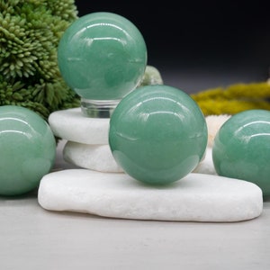 Pode incluir: Quatro esferas lisas de jade verde, cada uma com um acabamento polido, são exibidas em  apoios de pedra branca. As esferas estão dispostas em forma de pirâmide, com a esfera maior no topo.