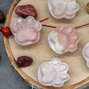 May include: Five pink and white paw print shaped stones on a wooden surface. The stones are polished and have a speckled appearance.