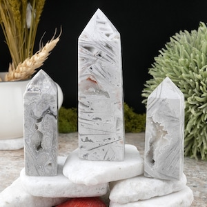 Op de afbeelding: Drie grijze en witte agaat kristallen torens van verschillende groottes. De kristallen hebben een gepolijst oppervlak met unieke patronen en een puntige top. Ze worden getoond op witte stenen, met groen en tarwearen op de achtergrond.