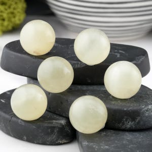 May include: Six pale green, spherical stones are arranged on dark gray, rectangular stones. The spheres have a smooth, polished appearance. The background includes a blurred view of a white and gray striped bowl and green moss.