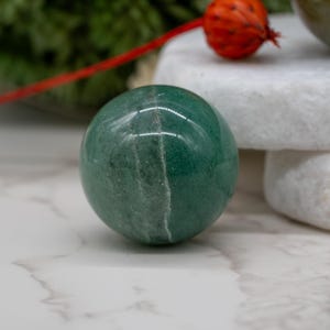 May include: A smooth, green aventurine sphere with a white streak running through the center. The sphere is resting on a white surface.