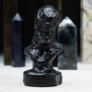 May include: A black sculpture of a bust with a detailed, textured design. The bust is in the foreground, with several crystal obelisks of varying colors and patterns in the background. The sculpture has a dark, glossy finish.
