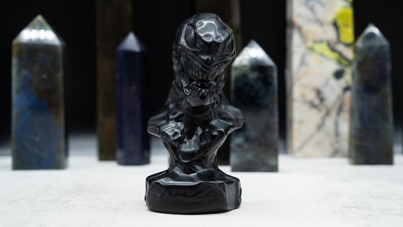 May include: A black sculpture of a bust with a detailed, textured design. The bust is in the foreground, with several crystal obelisks of varying colors and patterns in the background. The sculpture has a dark, glossy finish.