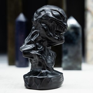 May include: A black sculpture of a creature with a detailed, textured surface. The sculpture has a menacing expression with visible teeth and a defined jawline. Various crystal obelisks are in the background.