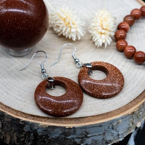 May include: A pair of brown sandstone earrings with silver hooks. The earrings are round with a hole in the center. The earrings are on a wooden surface.