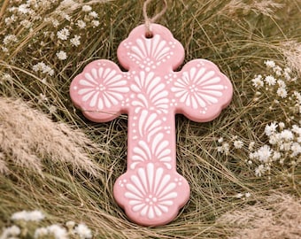 May include: A pink cross-shaped ornament with white floral designs. The cross is hanging by a natural twine loop. The background features dried grass and small white flowers, creating a rustic and decorative aesthetic.