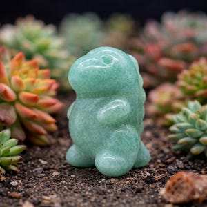 Op de afbeelding: Een kleine, lichtgroene dinosaurusfiguur van edelsteen. De dinosaurus is gesneden met afgeronde kenmerken en een getextureerd oppervlak. Het beeldje staat tegen een achtergrond van kleurrijke vetplanten en donkere aarde.
