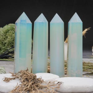 May include: Four opalescent crystal obelisks with pointed tops. The crystals have a soft, pearly white color with hints of blue and green. They are arranged on white stones with dried foliage in the foreground, creating a natural, earthy aesthetic.