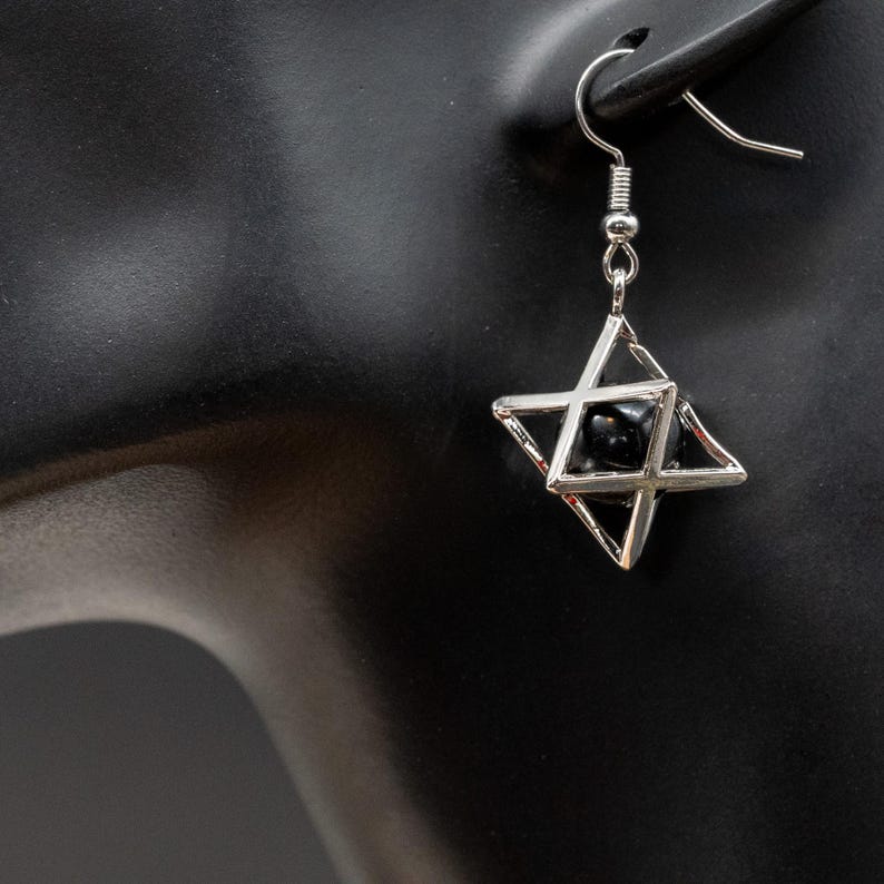 May include: A silver-colored earring featuring a geometric star design. The earring has a small black bead at the center of the star. The earring is hanging from a silver-colored hook.