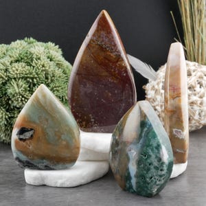 May include: Four polished gemstone sculptures in shades of brown, green, and cream. The stones are teardrop and obelisk shapes, displayed on white platforms. The background includes greenery and a dark surface.