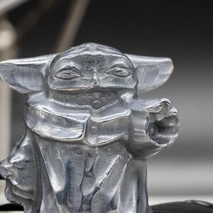 May include: A detailed, silver-colored figurine of a character with large ears and a pointed face, in a gesture as if using the Force. The figure is set against a blurred background with other dark objects.