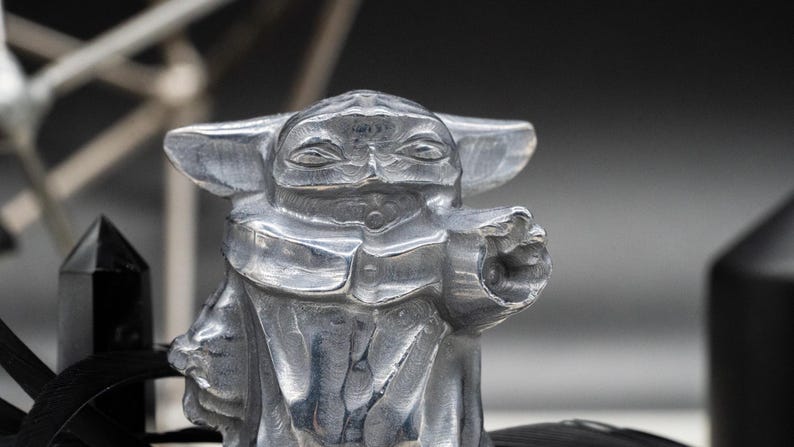 May include: A detailed, silver-colored figurine of a character with large ears and a pointed face, in a gesture as if using the Force. The figure is set against a blurred background with other dark objects.