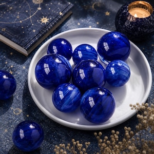 Peut inclure: Plusieurs sphères de cristal bleu foncé avec des motifs tourbillonnants blancs, disposées sur une assiette blanche. Les sphères, de tailles variées, ont un aspect brillant et reflètent la lumière. L'arrière-plan comprend une surface bleu foncé avec un motif céleste et une bougie allumée.