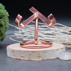 May include: A copper-coloured metal stand with a unique design, featuring two curved arms and a round base. The stand is displayed on a wooden surface.
