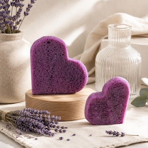 May include: Two heart-shaped, purple bath sponges. One sponge is on a small wooden stand. A bundle of lavender and a vase with lavender stems are in the background. A clear glass bottle is also in the background.