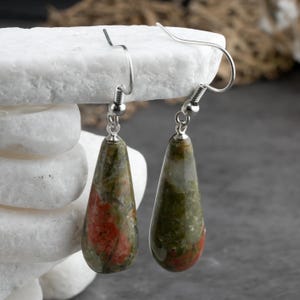 May include: A pair of drop earrings featuring polished, teardrop-shaped stones with a mottled pattern of olive green, forest green, and salmon pink. The stones are suspended from silver-toned metal fishhook wires with small spherical accents.