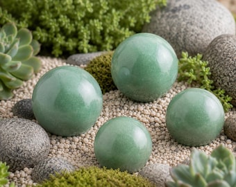 May include: Four smooth, polished, spherical green aventurine stones arranged among various natural grey river rocks, small light-colored pebbles, patches of green moss, and succulent plants, creating a balanced and textured composition.