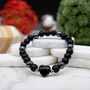 May include: Black beaded bracelet featuring matte onyx and hematite beads, with a heart-shaped onyx centerpiece. The bracelet is strung on an elastic band and has silver-colored accents. A stylish accessory for everyday wear.