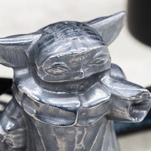 May include: A silver-colored figurine of a character with large ears and a robe. The figurine has a detailed, textured surface and is set against a blurred background with black accents.