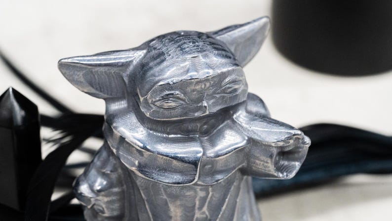 May include: A silver-colored figurine of a character with large ears and a robe. The figurine has a detailed, textured surface and is set against a blurred background with black accents.