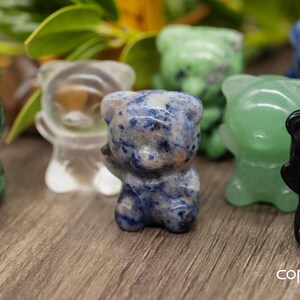 May include: A collection of seven small, carved bear figurines made of various gemstones. The bears are in different colors, including green, blue, clear, and black.