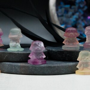 May include: A collection of colorful, translucent crystal figurines in various shades of purple, pink, green, and yellow. The figurines are displayed on gray stone platforms, with a blurred background featuring decorative elements.