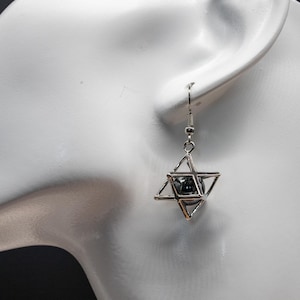 May include: A silver earring with a geometric star design. The star is made of interconnected triangles, with a dark, spherical bead at its centre. The earring hangs from a simple hook.