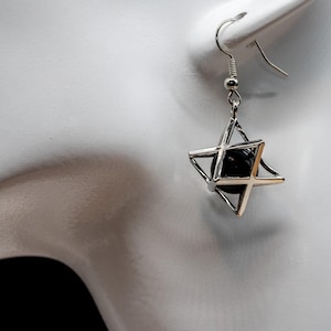 May include: Silver-tone metal earring featuring a geometric star design with a black sphere at the center. The earring has a hook closure and is displayed against a white background.