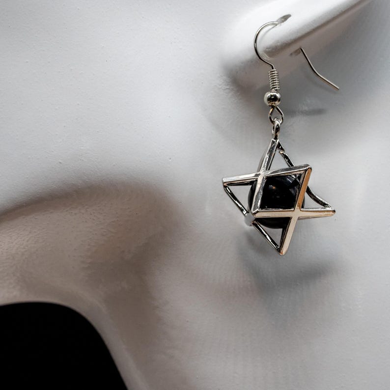 May include: Silver-tone metal earring featuring a geometric star design with a black sphere at the center. The earring has a hook closure and is displayed against a white background.