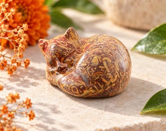 May include: A small, polished stone figurine of a curled-up sleeping cat. The surface features a complex, mottled pattern of reddish-brown, tan, and cream-colored fossilized inclusions throughout the smooth, rounded stone material.