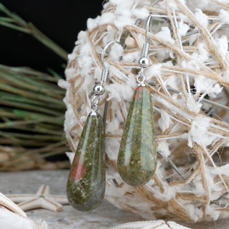 May include: A pair of teardrop-shaped earrings featuring polished green and red mottled stone pendants suspended from silver-toned metal fishhook wires. The smooth, rounded stones display natural variations in color and texture throughout.