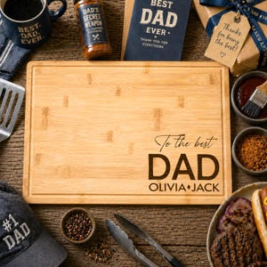 May include: A rectangular bamboo chopping board featuring a laser-engraved design that reads To the best DAD followed by the names OLIVIA and JACK. The board has a grooved perimeter and a light wood grain finish, positioned on a rustic wooden surface.