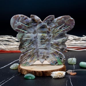 May include: A iridescent butterfly shaped sculpture made from mother of pearl. The sculpture is sitting on a wooden base and surrounded by green and orange stones.
