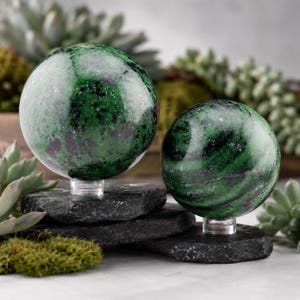May include: Two polished spherical stones featuring a mottled pattern of deep forest green, black, and vibrant magenta inclusions. Each sphere is mounted on a small, clear acrylic stand, resting atop a flat, dark gray, textured stone base.
