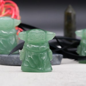 May include: Three green jadeite figurines of a character with large ears and a robe. The figurines are on a white surface with a dark background. Other objects include a white stone, a black ribbon, and a dark green crystal.