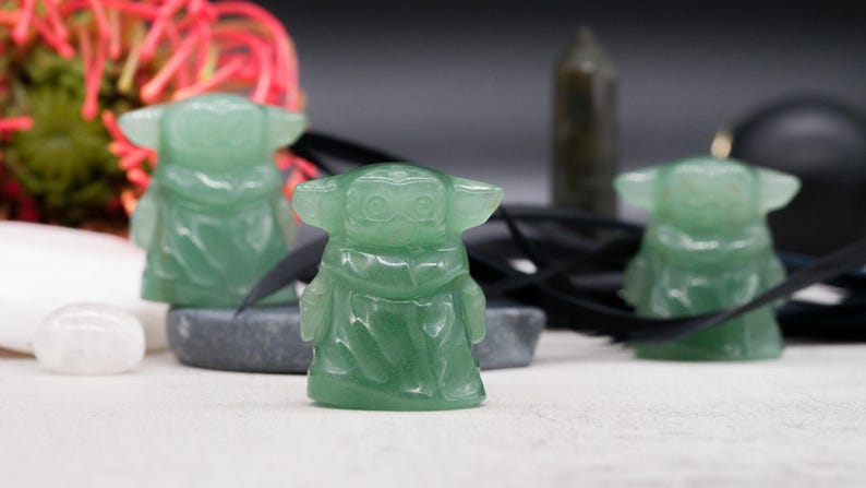 May include: Three green jadeite figurines of a character with large ears and a robe. The figurines are on a white surface with a dark background. Other objects include a white stone, a black ribbon, and a dark green crystal.