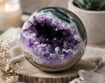 Könnte beinhalten: Eine kugelförmige Amethyst-Geode mit tiefviolettem Kristall-Innenraum. Die äußere Schale ist eine Mischung aus Grün und Grau, mit einem goldbraunen Sockel. Die Geode liegt auf einer Holzscheibe, mit einem klaren Bergkristall in der Nähe.