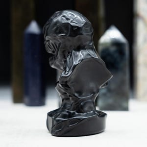 May include: A black sculpture of a bust with a textured surface. The bust is in the foreground, with various crystal towers in the background. The sculpture has a dark, glossy finish.