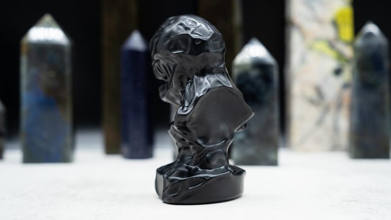 May include: A black sculpture of a bust with a textured surface. The bust is in the foreground, with various crystal towers in the background. The sculpture has a dark, glossy finish.