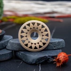 Puede incluir: Un expansor de oreja de madera con un diseño circular que presenta un patrón repetitivo de triángulos. El expansor es de color marrón claro y tiene un agujero en el centro.