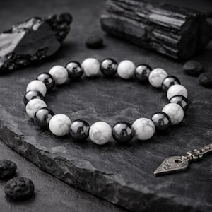 May include: A beaded bracelet featuring alternating white and dark gray spherical beads. The bracelet is displayed on a dark gray, textured surface, with additional dark gray stones and a decorative metal piece in the background. The bracelet is a jewelry item.