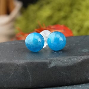 Op de afbeelding: Twee ronde, helderblauwe edelsteen oorbellen. De oorbellen hebben een levendige turkooizen kleur en lijken gepolijst. Ze liggen op een donkere, getextureerde ondergrond, met een vage groene en rode achtergrond.